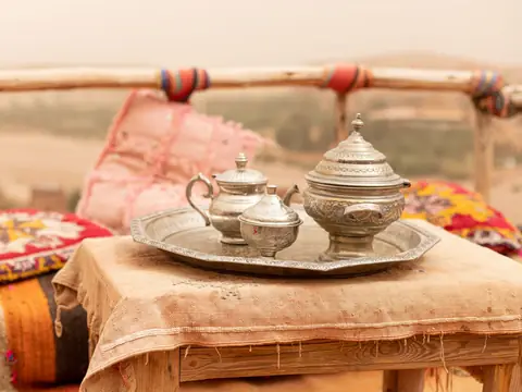 A silver teapot, sugar bowl and covered dish on a round tray, on a low table with bright cushions