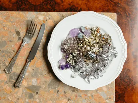 A white plate heaped with odd earrings, chains, beads and broken pieces of jewelry beside an old fork and knife