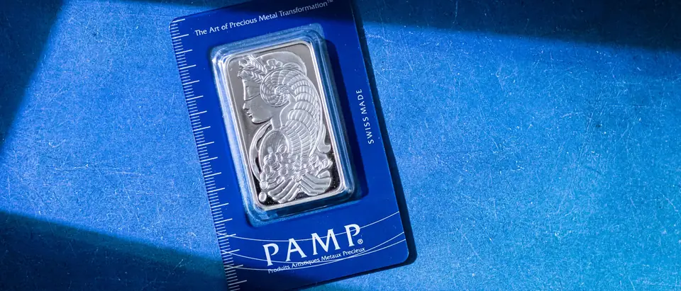 A fine silver bar in its sealed assay card lying in a band of sunlight on blue cloth