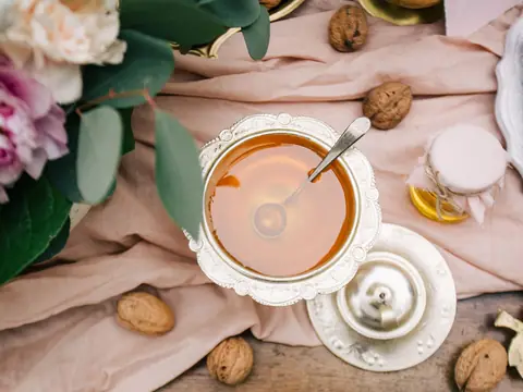 A silver bowl of honey with a spoon and a small covered silver pot on pink linen