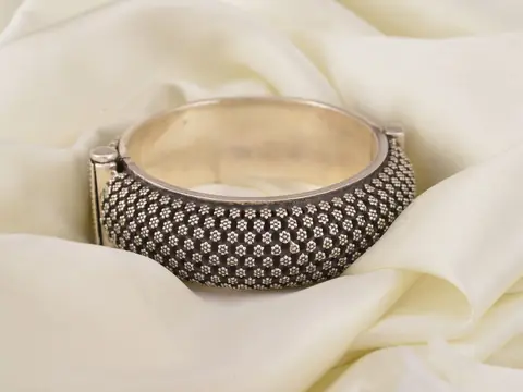 A wide silver cuff bracelet patterned with small flowers, resting in cream cloth