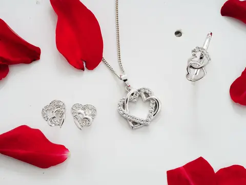 A silver heart pendant on a chain and a pair of heart earrings on white, with red petals
