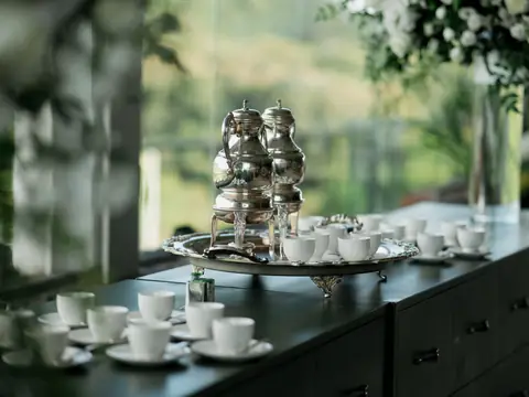 A silver coffee and tea service on a large tray, with white cups set out along a dark sideboard